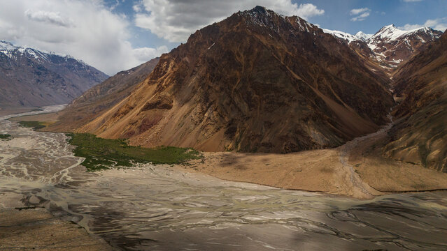 Aerial view of the rugged Bartang Valley featuring a wide braided riverbed, steep rocky mountains with snow-capped peaks, and green vegetation in Chah Ab, Gorno-Badakhshan Auto, Tajikistan.