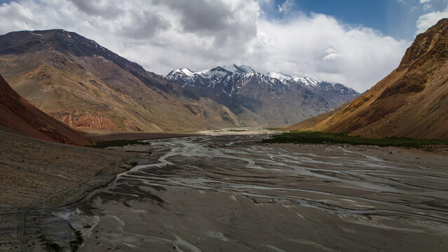 Aerial view of the braided Bartang River flowing through a rugged mountain valley with snow-capped peaks under a cloudy sky in Chah Ab, Gorno-Badakhshan Auto, Tajikistan.