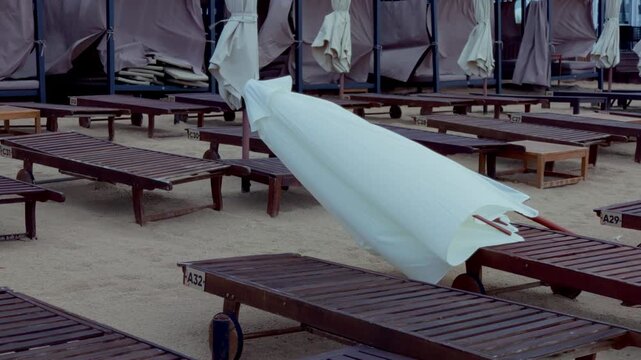 Close up tracking shot showing collapsed sunshade umbrella lying over sand beside empty lounge chairs on Baltic coastline in Jurmala during windy day.