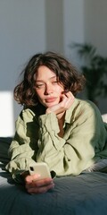 Obraz premium Young woman holding smartphone and gazing in warm sunlight for lifestyle background and candid indoor portrait visuals