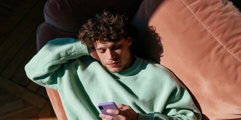 Obraz premium Young man lying on terracotta couch checking smartphone with arm behind head in mint sweater warm window light for social media lifestyle visual