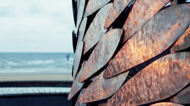 Close up tracking view displays metallic scales and copper textures of public art installation with blurred Baltic Sea waves and sandy beach in background