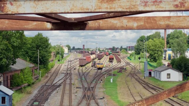 Elevated static shot through rusty bridge girders overlooking railway yard with cargo carriages, industrial depot, and junction switches under overcast sky in Daugavpils.