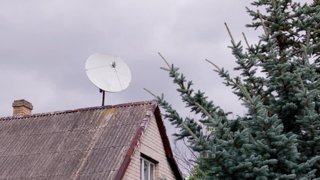 Close up shot of white parabolic disk receiver mounted on weathered roof beside branches of pine tree under cloudy sky, depicting private media access in Baltic region.