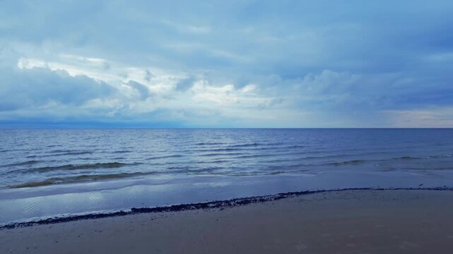 Static wide shot captures tranquil Baltic Sea surf lapping onto wet sand in Jurmala Latvia under cloudy summer evening sky offering serene coastal view.
