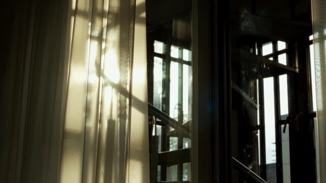 Warm evening sunlight illuminates sheer curtain hanging beside modern window frame, casting gentle shadows across tranquil room interior during golden hour in static closeup.