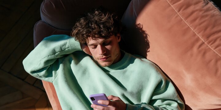 Young man lying on terracotta couch checking smartphone with arm behind head in mint sweater warm window light for social media lifestyle visual