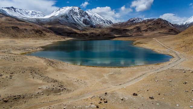 Aerial view of a turquoise alpine lake in the Pamir Mountains, with snow-capped peaks, arid terrain, and a dirt road in Jirgatol, Gorno-Badakhshan Auto, Tajikistan.