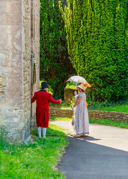 A man in a Victorian red coat gestures to welcome a woman in a period floral dress and parasol towards a historic building, participating in a historical reenactment.