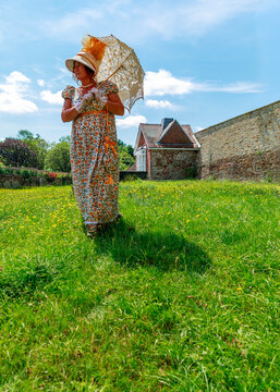 A woman dressed in vintage Regency-style clothing and holding a delicate parasol walks gracefully through a sunlit field, participating in a historical reenactment or themed garden photoshoot.