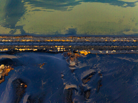 Aerial view of a straight asphalt road cutting through a dark volcanic landscape and shallow coastal waters in Iceland.