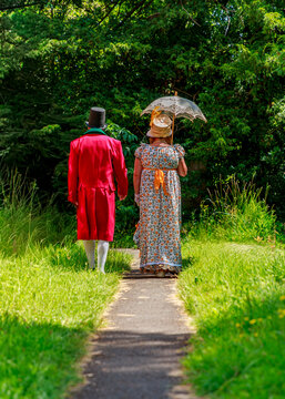 A man in a historical red tailcoat and a woman in a period floral dress stroll along a garden path, likely participating in a Regency or Victorian-era costume reenactment event in nature.