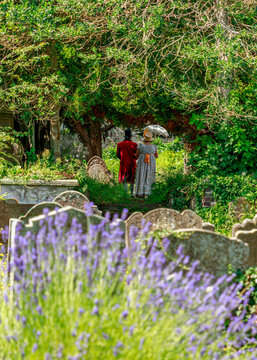 A couple dressed in Victorian-era attire walk through a lush, historic cemetery, likely participating in a period re-enactment or paying respects at a memorial event amidst the gravestones.