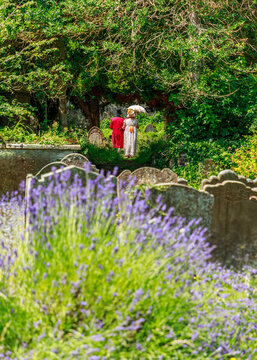 A couple dressed in 19th-century Victorian attire walks through a serene, flower-filled cemetery, creating a nostalgic scene that evokes a historical memorial or a themed garden stroll.