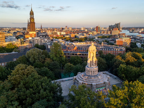 Aerial view of the Bismarck Monument surrounded by trees with St. Michaeliskirche and the Hamburg cityscape in the background during golden hour Hamburg, Germany.