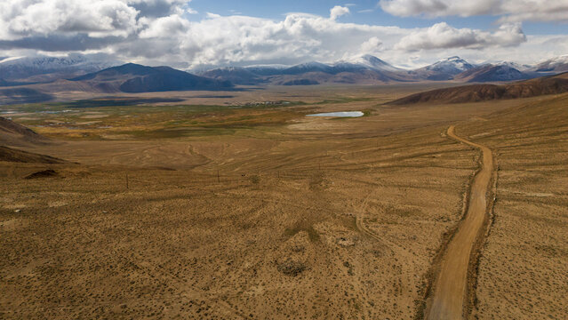 Aerial view of the arid landscape near Bulunkul lake, with a winding dirt road cutting through dry terrain and snow-capped mountains in Chah Ab, Gorno-Badakhshan, Tajikistan.