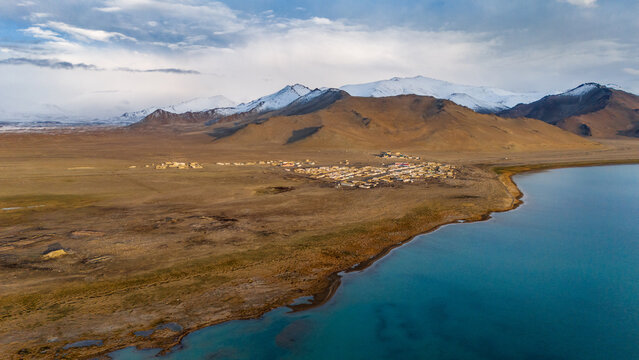 Aerial view of Karakul Lake with its turquoise waters, surrounded by arid plains and snow-capped mountains under a cloudy sky in Chah Ab, Gorno-Badakhshan Auto, Tajikistan.