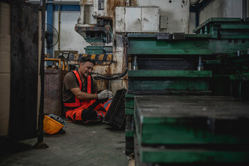 Fototapeta premium Injured male industrial worker sitting in pain on the floor holding his knee after a workplace accident with a hard hat on the ground inside a manufacturing factory.
