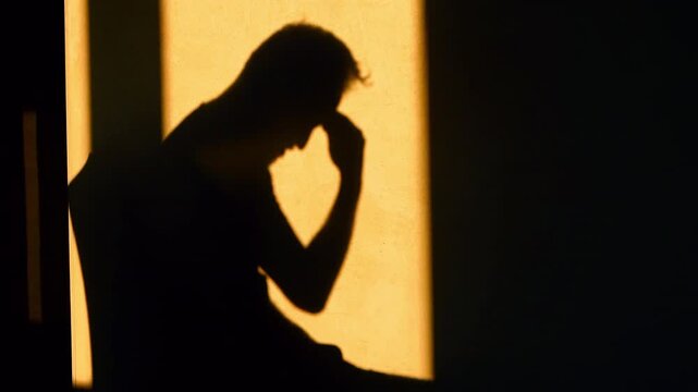 Shadow silhouette of a depressed man on orange background. Sitting on a chair with head in hands and rubbing temples in pain from a headache