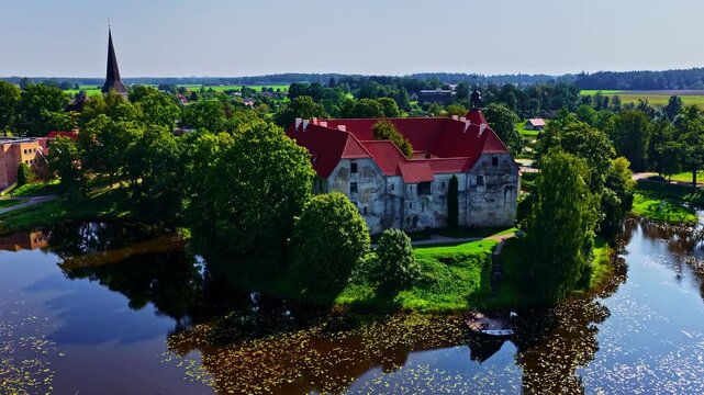 Aerial view of the historic Jaunpils Castle with its distinctive red roof and stone walls, surrounded by a moat and lush greenery in Jaunpils, Tukums Municipality, Latvia.