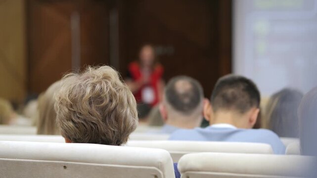 Audience members sit facing forward, listening attentively in a conference room. Focus on backs of heads, showing engagement and concentration. Blurred presenter in background adds depth