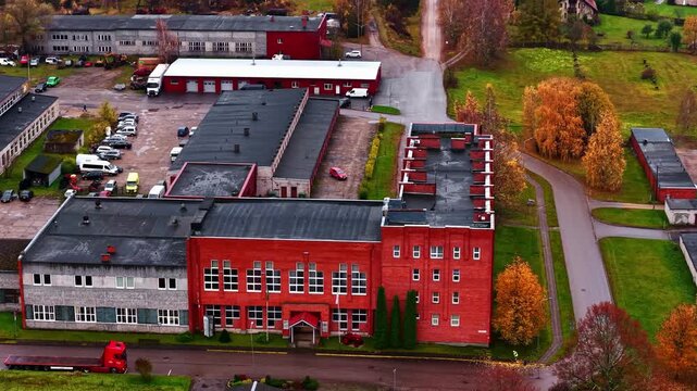 Aerial view of industrial buildings, warehouses, and a parking lot with various vehicles in Priekuli, Cesis Municipality, Latvia.