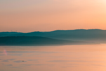 Fototapeta premium Tranquil panoramic view of a calm sea bay reflecting a warm golden sunset sky with silhouetted misty mountains.