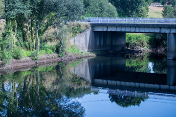 Fototapeta premium A concrete road bridge over a calm river reflecting symmetrically in the still water surrounded by green trees.