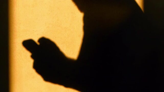 Close-up hands using a phone. Casting silhouette shadow on the yellow wall