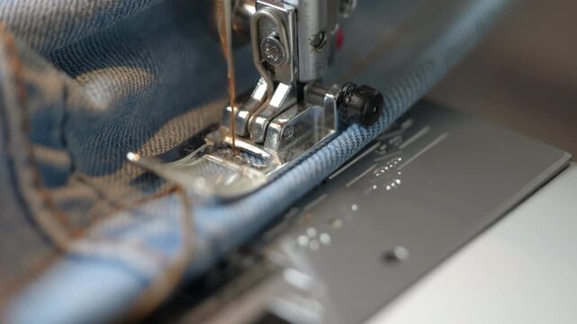 Close-up of sewing machine stitching denim fabric. Extreme close-up shot of a sewing machine's needle and presser foot stitching a seam on blue denim jeans fabric, illustrating tailoring