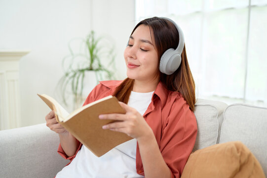 Happy Asian Woman Reading a Book While Wearing Wireless Headphones and Relaxing at Home