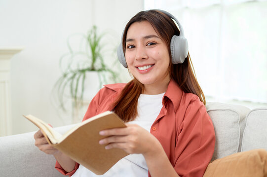 Happy Asian Woman Reading a Book While Wearing Wireless Headphones and Relaxing at Home