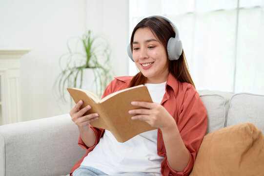 Happy Asian Woman Reading a Book While Wearing Wireless Headphones and Relaxing at Home