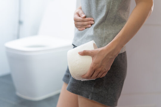 Stomach ache, sick asian young woman holding stomach pain and holding tissue paper roll in bathroom, standing near toilet, girl suffer from diarrhea her stomach painful. Hygiene, health care concept.