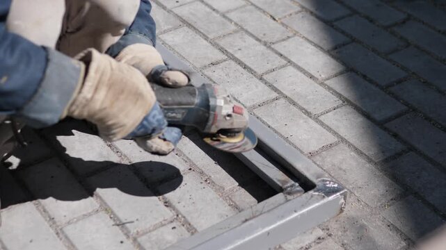 Gloved worker cuts metal rail inside light gray brick paving with handheld grinder, small sparks at contact point, strong sunlight, hard shadows, dusty surface, close elevated viewpoint, practical