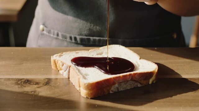 Pouring dark apron cook tilting steel pitcher at counter, coating white bread slice with syrup