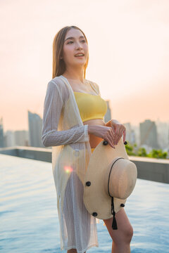 Playful joyful young woman laughing with closed eyes holding straw hat at rooftop pool sunset, enjoying vacation bliss, freedom, luxury leisure, wellbeing, and cheerful vitality.