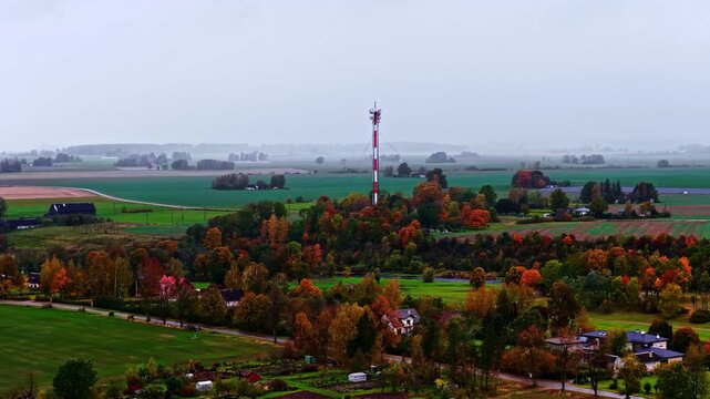 Aerial view of a tall communication tower rising above vibrant autumn trees and sprawling agricultural fields in Bauska, Bauska Municipality, Latvia.