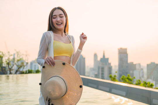 Playful joyful young woman laughing with closed eyes holding straw hat at rooftop pool sunset, enjoying vacation bliss, freedom, luxury leisure, wellbeing, and cheerful vitality.