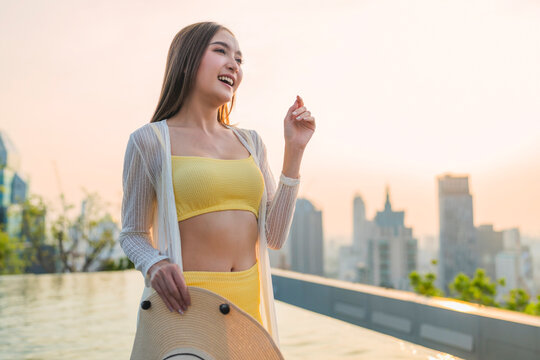 Playful joyful young woman laughing with closed eyes holding straw hat at rooftop pool sunset, enjoying vacation bliss, freedom, luxury leisure, wellbeing, and cheerful vitality.