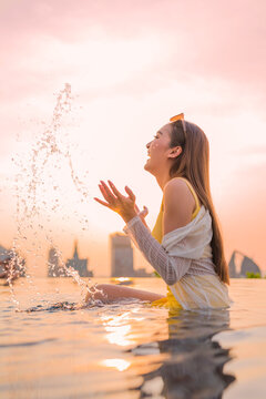 Carefree young woman enjoying summer vacation freedom, splashing water at rooftop infinity pool sunset, expressing pure joy, happiness, vitality, and luxury lifestyle retreat.