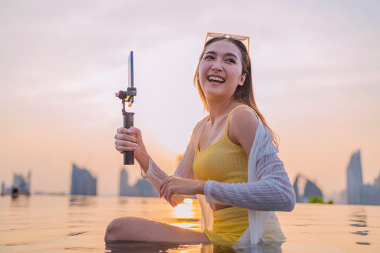 Cheerful young Asian woman vlogging at sunset by a rooftop infinity pool, laughing while recording content on her smartphone gimbal against a glowing city skyline, living the carefree travel lifestyle