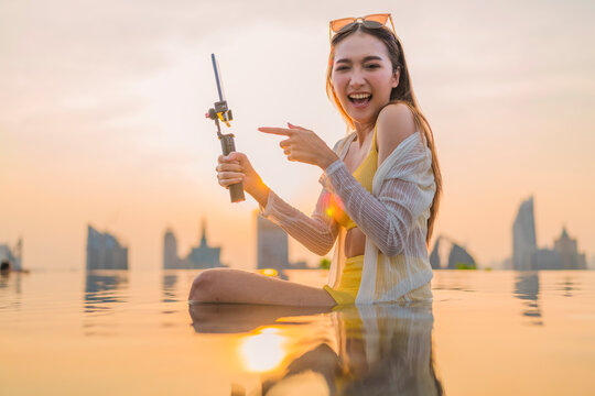 Cheerful young Asian woman vlogging at sunset by a rooftop infinity pool, laughing while recording content on her smartphone gimbal against a glowing city skyline, living the carefree travel lifestyle