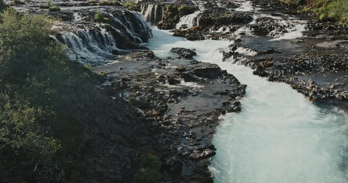Br&uacute;arfoss waterfall cascading through dark volcanic rocks in Iceland, with vivid turquoise glacial water flowing through a rugged natural landscape surrounded by lush green vegetation.