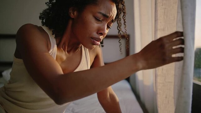 Gasping then coughing woman wearing tank top, pants on bed, seeking fresh air, peering out window