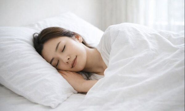 白いベッドで布団に包まれ穏やかに眠る若い女性の朝のリラックスタイム