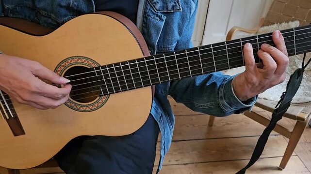 A close-up of a musician s hands playing an acoustic classical guitar at home, focusing on finger placement, the wooden floor, denim jacket, and the intimate atmosphere of practice and concentration.
