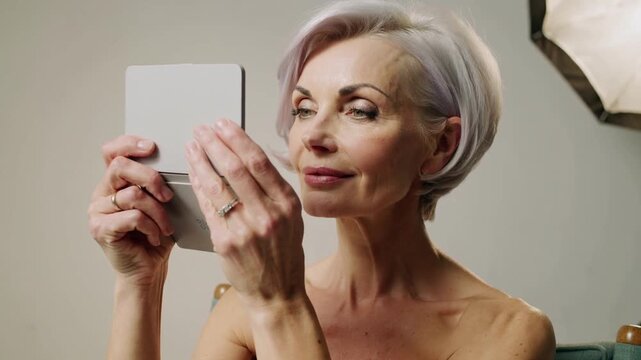 Lifting mirror up, senior woman adjusting, checking makeup in studio with softbox in strapless top