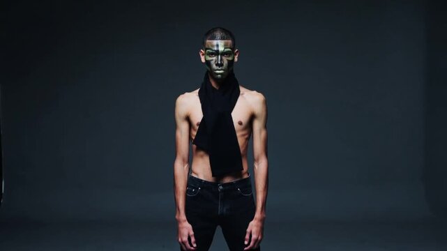 Raising shirtless model placing long black scarf around neck in studio, highlighting painted face