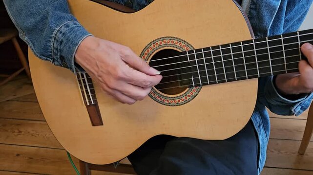 A close-up of a musician s hands playing an acoustic classical guitar at home, focusing on finger placement, the wooden floor, denim jacket, and the intimate atmosphere of practice and concentration.